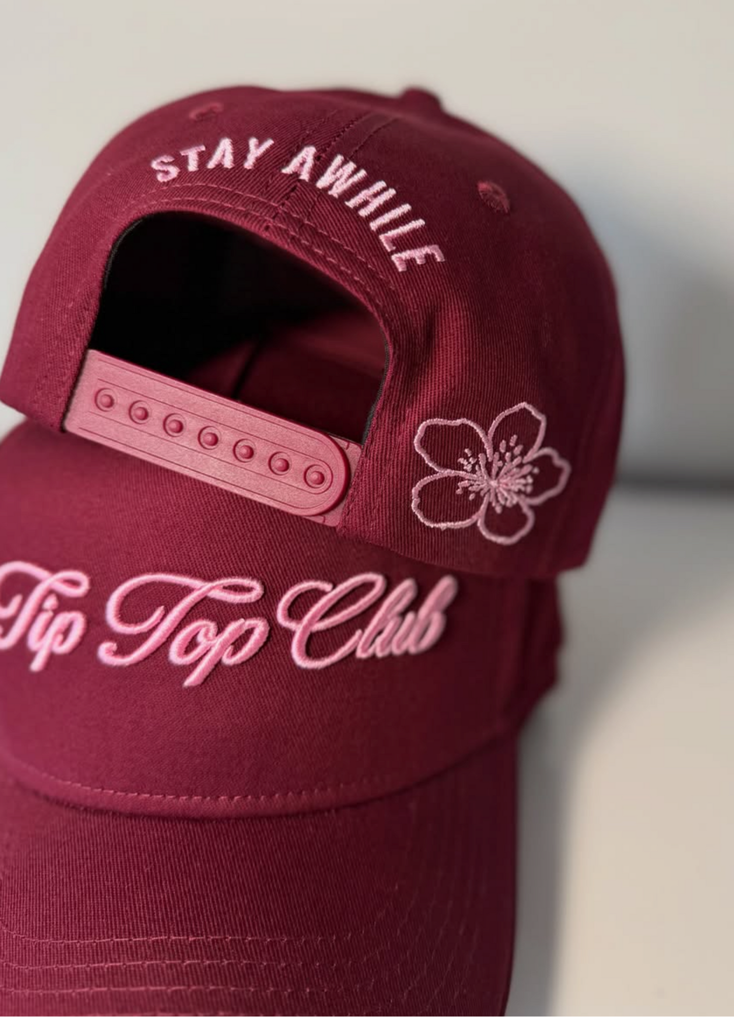 A close-up of a burgundy baseball cap with pink embroidered text reading 'Stay Awhile' on the back and 'Tip Top Club' on the brim, plus a pink flower outline on the side.